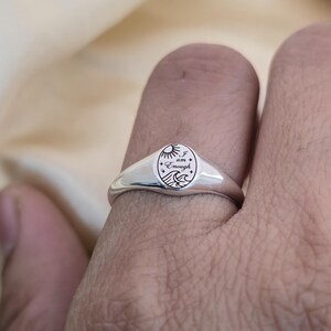 May include: A silver signet ring with an oval face. The face features an engraved design of a sun, waves, and the words "I am Enough". The ring is worn on a finger.