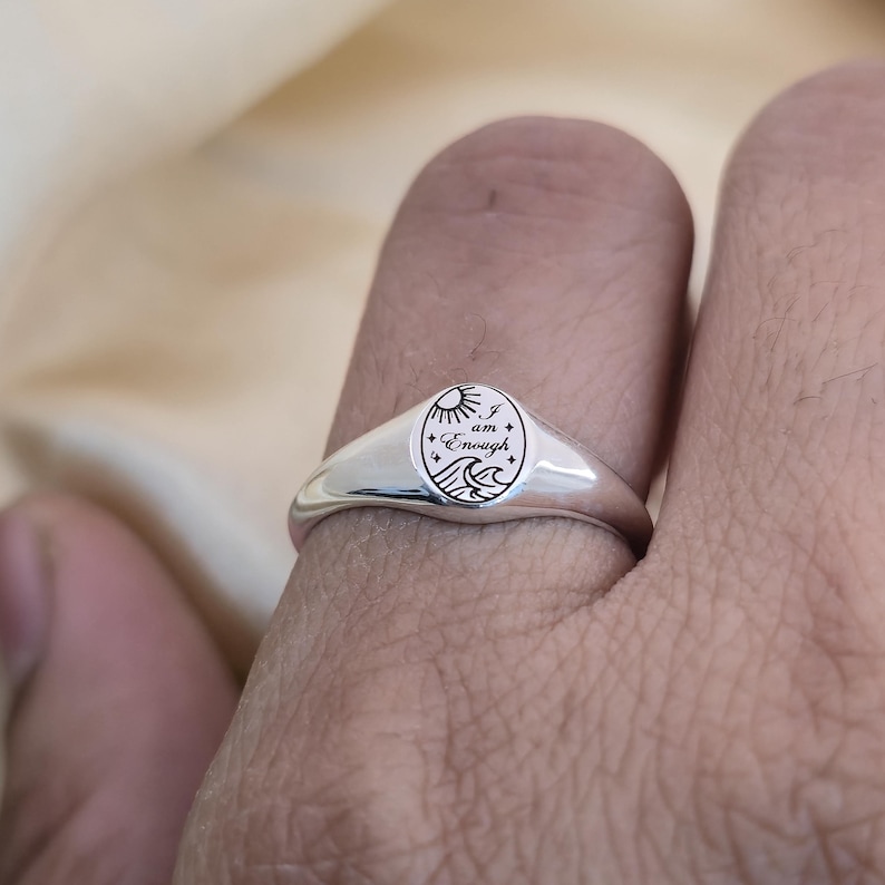 May include: A silver signet ring with an oval face. The face features an engraved design of a sun, waves, and the words "I am Enough". The ring is worn on a finger.