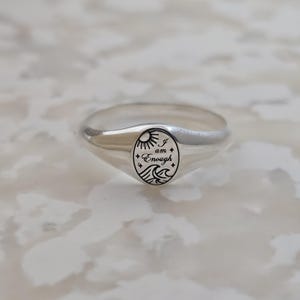 May include: A silver signet ring with an oval face. The face features a sun, waves, and the words "I am Enough" in a black, stylized font. The ring has a polished finish and a smooth band.