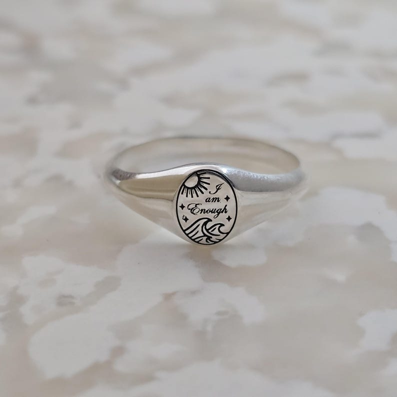 May include: A silver signet ring with an oval face. The face features a sun, waves, and the words "I am Enough" in a black, stylized font. The ring has a polished finish and a smooth band.