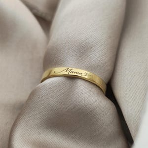 May include: A gold ring engraved with the word "Mama" in cursive, with a heart symbol. The ring is set against a backdrop of a soft, neutral-toned fabric. The design is simple and elegant, suitable for everyday wear.