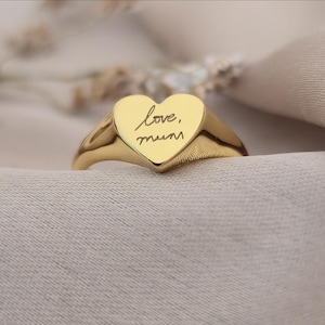Peut inclure: Une bague dorée avec une face en forme de cœur. Le cœur porte l'inscription manuscrite "love, mum". La bague est mise en valeur sur un fond neutre, soulignant son design.