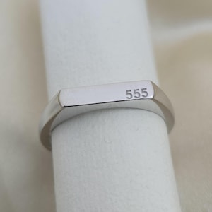 Puede incluir: Un anillo de sello plateado con el n&uacute;mero "555" grabado en la cara rectangular. El anillo se muestra sobre un fondo blanco, resaltando su superficie pulida y reflectante.