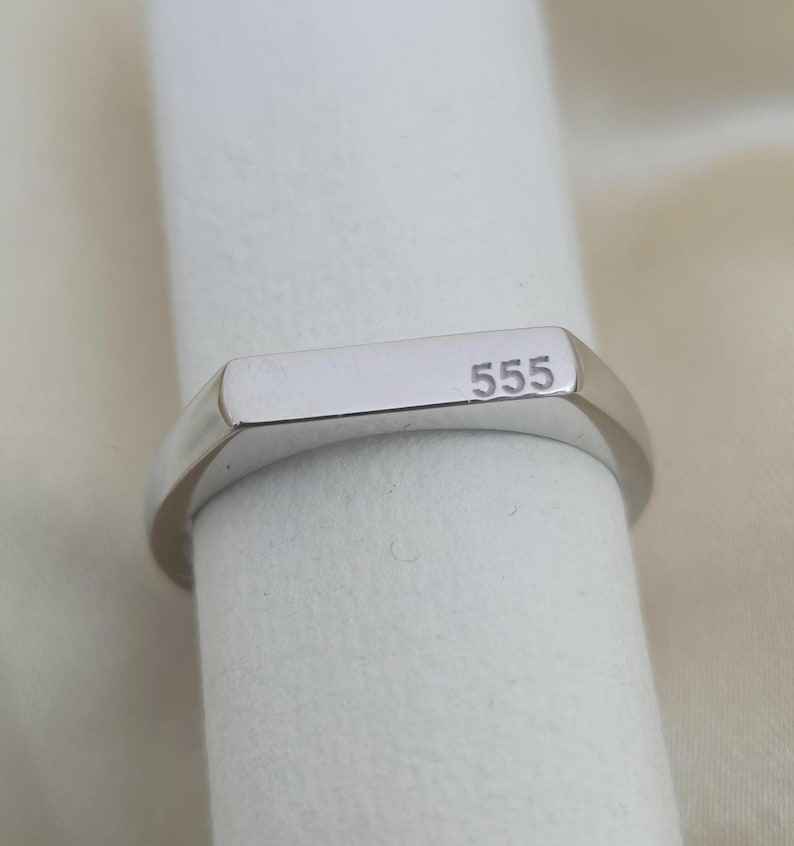 Puede incluir: Un anillo de sello plateado con el n&uacute;mero "555" grabado en la cara rectangular. El anillo se muestra sobre un fondo blanco, resaltando su superficie pulida y reflectante.