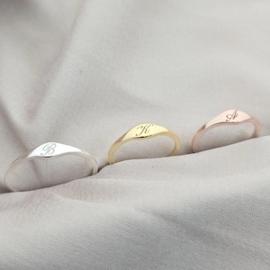 May include: Three signet rings in silver, gold, and rose gold, each engraved with a single initial. The rings are displayed on a soft, neutral-toned fabric. The initials are "B", "K", and "A".