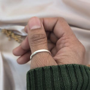Puede incluir: Un anillo plateado con una banda lisa y redondeada. El anillo se muestra en el pulgar de una persona, con un su&eacute;ter verde en primer plano y un fondo de tonos neutros.