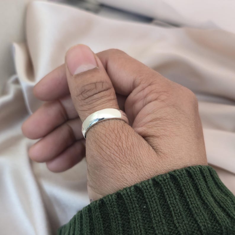 Puede incluir: Un anillo plateado con una superficie lisa y pulida se lleva en el pulgar de una persona. El anillo refleja la luz, creando una apariencia brillante y brillante. La persona lleva un su&eacute;ter verde.