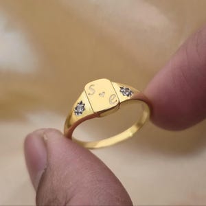 Puede incluir: Un anillo de sello dorado con una cara rectangular. La cara tiene grabadas las letras "S" y "E". Pequeñas piedras decorativas están engastadas a cada lado de la cara. El anillo está sujeto por los dedos de una persona.