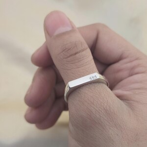 Puede incluir: Un anillo plateado con una parte superior rectangular que presenta el n&uacute;mero "555" grabado. El anillo se lleva en un dedo, mostrando su dise&ntilde;o simple y moderno. La superficie pulida del anillo refleja la luz, resaltando su brillo met&aacute;lico.
