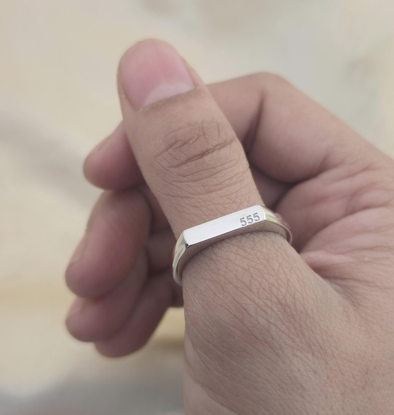 Puede incluir: Un anillo plateado con una parte superior rectangular que presenta el n&uacute;mero "555" grabado. El anillo se lleva en un dedo, mostrando su dise&ntilde;o simple y moderno. La superficie pulida del anillo refleja la luz, resaltando su brillo met&aacute;lico.