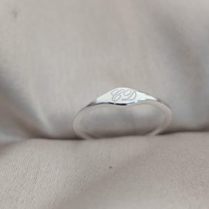 Op de afbeelding: Een gepolijste zilveren zegelring met een glad, afgerond oppervlak gegraveerd met de cursieve letters CD. De ringband is slank en loopt taps toe naar de voorzijde, met een reflecterende metalen afwerking en een strak, eenvoudig ontwerp.