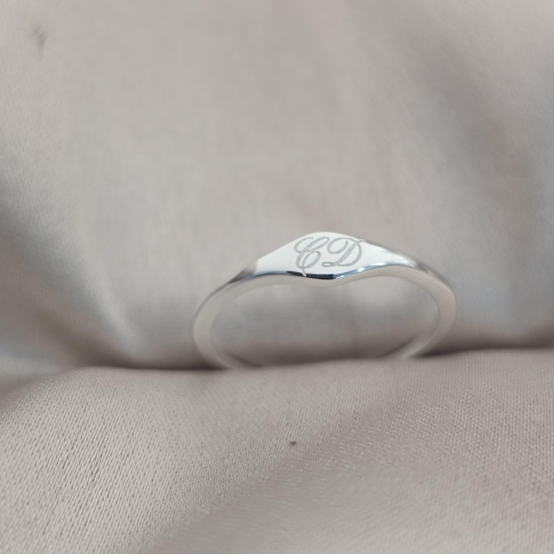Op de afbeelding: Een gepolijste zilveren zegelring met een glad, afgerond oppervlak gegraveerd met de cursieve letters CD. De ringband is slank en loopt taps toe naar de voorzijde, met een reflecterende metalen afwerking en een strak, eenvoudig ontwerp.