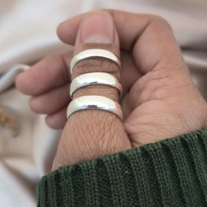 Puede incluir: Tres anillos de plata apilados en un dedo. Los anillos tienen una superficie lisa y pulida y una banda redondeada. El fondo es de un color suave y neutro, y la persona lleva un jersey verde.