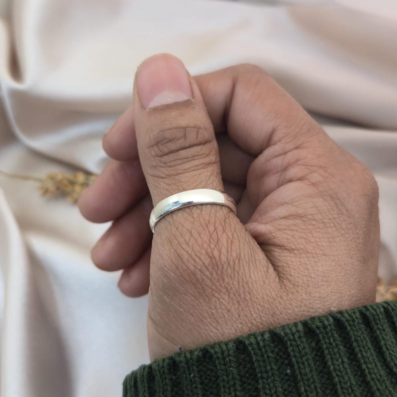 Puede incluir: Un anillo plateado brillante se lleva en el pulgar. El anillo tiene un dise&ntilde;o liso y redondeado y refleja la luz. La persona lleva un su&eacute;ter verde. El fondo es de color claro y neutro.