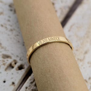 Op de afbeelding: Een goudkleurige bandring met de gegraveerde Romeinse cijfers VI.XII.MMXX. De ring wordt getoond op een neutrale ondergrond, waarbij de gepolijste afwerking en het eenvoudige ontwerp worden benadrukt. De inscriptie van de ring suggereert een datum.