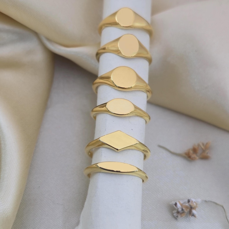 Puede incluir: Una colecci&oacute;n de anillos de sello dorados con varias formas, incluyendo &oacute;valos, redondos, diamantes y oblongos. Los anillos se exhiben sobre una superficie cil&iacute;ndrica blanca contra un fondo suave y neutro. Los anillos son brillantes y reflectantes.
