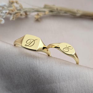以下が含まれることがあります： 2つの金のシグネットリング。1つのリングは、筆記体で「D」の文字が入った正方形の面を持っています。もう1つのリングは、筆記体で「E」の文字が入った楕円形の面を持っています。リングは薄いピンク色の生地の上にあります。