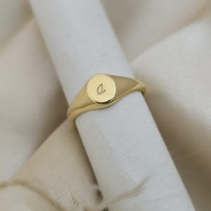 May include: A gold signet ring with a polished finish. The ring has a circular face with the letter "a" engraved. The ring is displayed on a white fabric background, highlighting its simple elegance.
