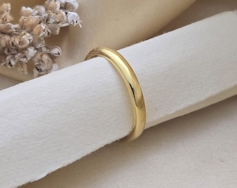May include: A gold wedding band is displayed on a white surface. The ring has a smooth, polished finish and a classic, rounded design. The band is a warm, yellow gold color, reflecting light. The background is a soft, neutral tone, highlighting the ring's details.