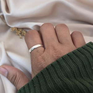 Puede incluir: Un anillo plateado con una banda lisa y redondeada se lleva en un dedo. El anillo refleja la luz, apareciendo brillante contra la piel. El fondo es una tela suave y neutra, y se ve una manga verde de un su&eacute;ter.