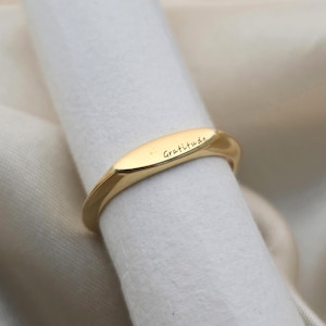 以下が含まれることがあります： 「Gratitude」の文字が刻まれたゴールドカラーのリング。リングは楕円形で、磨かれた金属製に見えます。白い表面にリングが表示されています。