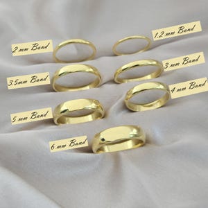 Könnte beinhalten: Eine Auswahl goldfarbener Ringe mit unterschiedlichen Bandbreiten, von 1,2 mm bis 6 mm, auf einem neutralen Stoff. Jeder Ring ist mit seiner Bandgröße in Millimetern beschriftet.