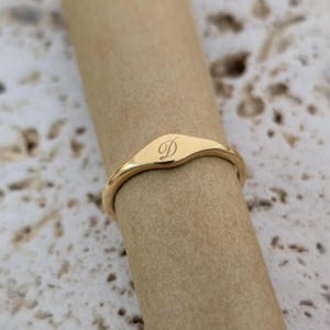 Puede incluir: Un anillo de sello dorado con un acabado liso y pulido. El anillo presenta una cara rectangular ligeramente curvada con la letra "D" grabada en una escritura delicada. El anillo se muestra en un dedo.
