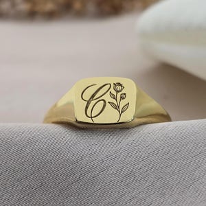 Könnte beinhalten: Ein goldfarbener Siegelring mit quadratischer Oberfläche. Auf der Oberfläche des Rings ist der Buchstabe "C" in Schreibschrift zusammen mit einem floralen Design abgebildet. Der Ring ist vor einem neutralen Hintergrund platziert.