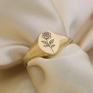May include: A gold-colored signet ring with an oval face. The ring features a detailed engraving of a rose with leaves. The ring is set against a soft, cream-colored fabric, highlighting its polished finish.