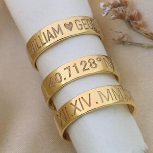 May include: Three gold-colored rings with engraved text. The top ring reads "WILLIAM ♥ GEORGE". The middle ring displays "40.7128° N". The bottom ring shows "XII.XIV.MMXX". The rings are stacked on a white surface.