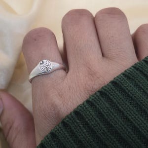 May include: A silver signet ring with a circular face featuring a sun, waves, and the text "I am Enough." The ring is worn on a finger, with a green sweater in the background.
