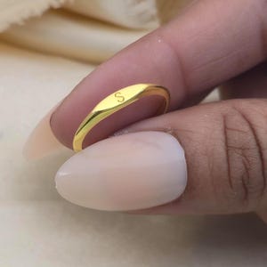 Puede incluir: Un anillo de sello dorado con la letra "S" grabada en la cara ovalada. El anillo tiene un acabado liso y pulido y un diseño simple y elegante. El anillo se sostiene cerca de la cámara.