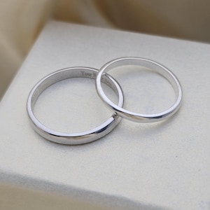 Peut inclure: Deux alliances en argent, l'une chevauchant l'autre. Les bagues sont lisses et brillantes, avec un design simple et classique. Une bague porte l'inscription "Your Name Here" gravée à l'intérieur. Les bagues sont posées sur un fond doux et neutre.