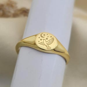 Op de afbeelding: Een gouden zegelring met een ronde voorkant met een gegraveerd ontwerp van een duif, een maan en sterren. De ring heeft een gepolijste afwerking en een gladde band. Een klassiek sieraad.