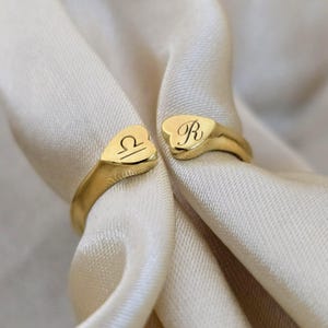 Op de afbeelding: Goudkleurige open ring met hartvormige uiteinden. Eén uiteinde heeft het Weegschaal-symbool, de andere de letter "R". De ring wordt getoond op een crèmekleurige stof, wat het ontwerp benadrukt.
