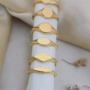 Puede incluir: Una colecci&oacute;n de anillos de sello dorados con varias formas, incluyendo ovaladas, redondas y de diamante. Los anillos se muestran sobre un objeto cil&iacute;ndrico blanco contra un fondo suave y neutro. Los anillos probablemente son de metal y tienen un acabado pulido.