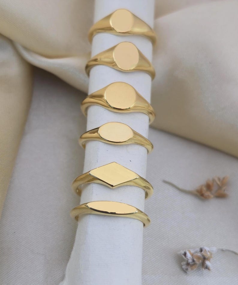 Puede incluir: Una colecci&oacute;n de anillos de sello dorados con varias formas, incluyendo ovaladas, redondas y de diamante. Los anillos se muestran sobre un objeto cil&iacute;ndrico blanco contra un fondo suave y neutro. Los anillos probablemente son de metal y tienen un acabado pulido.