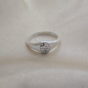 May include: A silver ring with a rounded band and a circular face. The face features a sun, waves, and the words "I am Enough" in a script font. The ring is displayed on a cream-colored fabric.