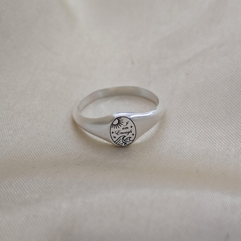 May include: A silver ring with a rounded band and a circular face. The face features a sun, waves, and the words "I am Enough" in a script font. The ring is displayed on a cream-colored fabric.