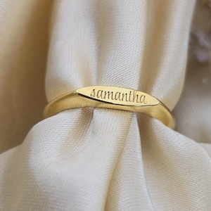 Puede incluir: Un anillo dorado con el nombre "Samantha" grabado en una placa rectangular. El anillo se presenta sobre un fondo de tela suave y blanquecina, resaltando la superficie pulida y el diseño elegante del anillo.
