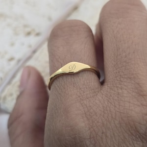 Op de afbeelding: Een gepolijste goudkleurige zegelring met een glad, plat, ovaalvormig oppervlak. In het midden van de ring is een enkele, elegante, cursieve hoofdletter D gegraveerd. De ringband is dun, afgerond en heeft een consistente gouden afwerking.