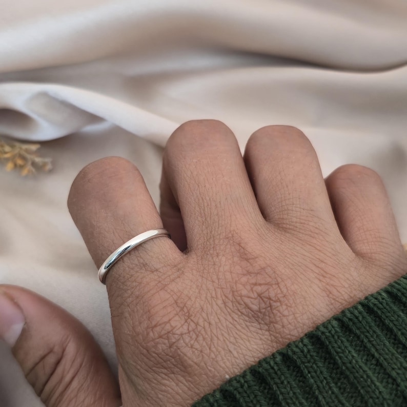 Puede incluir: Un anillo de plata sencillo y pulido se lleva en un dedo. El anillo tiene una banda lisa y redondeada. El fondo es de un color suave y neutro, con una manga verde visible en el encuadre. Una pieza de joyer&iacute;a cl&aacute;sica.