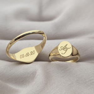 May include: Two gold signet rings. One ring has the engraved date "08-12-20". The other ring has the engraved initials "AJ" in a script font. Both rings are displayed on a neutral-colored fabric.