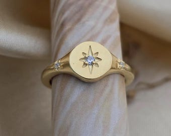 Puede incluir: Un anillo dorado con un diseño de estrella. El anillo tiene una cara redonda con un diseño de estrella y una pequeña piedra transparente en el centro. Dos piedras transparentes más pequeñas están engastadas en la banda.