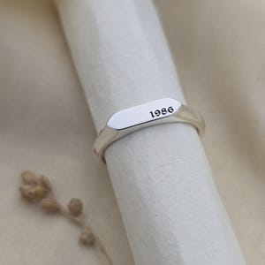 以下が含まれることがあります： 長方形の面を持つシルバーのシグネットリング。リングの表面には黒で「1986」の年号が刻まれています。リングは白い表面に表示され、柔らかくニュートラルなトーンの背景があります。