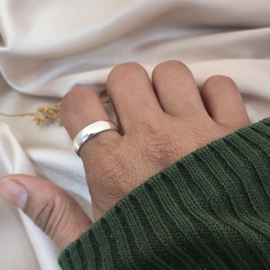 Puede incluir: Un anillo de plata pulida con una banda lisa y redondeada se lleva en un dedo. El anillo refleja la luz, creando una apariencia brillante y brillante. El anillo se coloca sobre un fondo de un su&eacute;ter verde y una tela color crema.