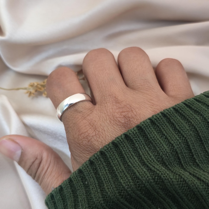 Puede incluir: Un anillo de plata pulida con una banda lisa y redondeada se lleva en un dedo. El anillo refleja la luz, creando una apariencia brillante y brillante. El anillo se coloca sobre un fondo de un su&eacute;ter verde y una tela color crema.