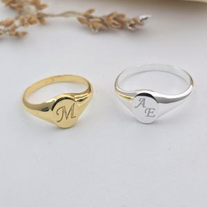 Puede incluir: Se muestran dos anillos de sello, uno dorado y otro plateado. El anillo dorado tiene la letra "M" grabada en su superficie. El anillo plateado tiene las letras "A" y "E". Ambos anillos tienen un acabado pulido.