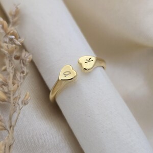 May include: A gold-coloured open ring featuring two heart-shaped terminals, each bearing a single engraved initial. The ring is presented against a white surface with a soft, neutral backdrop. Jewellery.