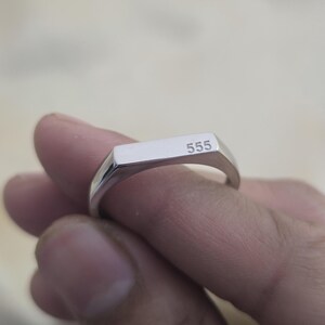 Puede incluir: Un anillo de sello plateado con el n&uacute;mero "555" grabado en la cara rectangular. El anillo est&aacute; sostenido por los dedos de una persona, mostrando su superficie pulida y reflectante. El anillo es una pieza de joyer&iacute;a simple y elegante.